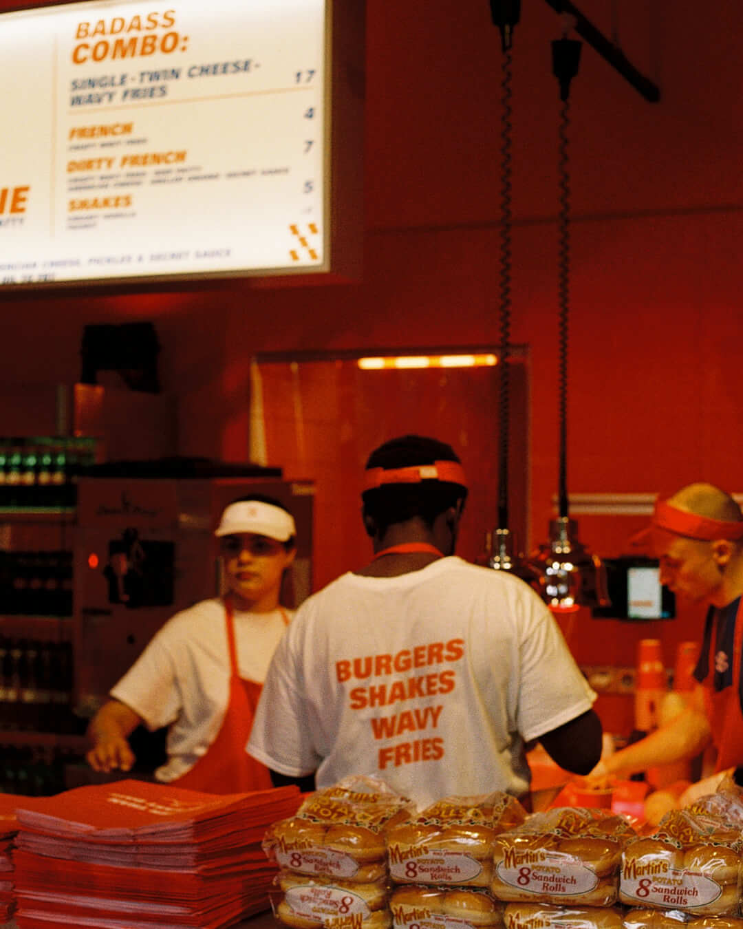 Inside the restaurant, staff wearing white shirts with red text reading "BURGERS SHAKES WAVY FRIES" work behind the counter. Visible items include packs of sandwich rolls, stacks of red trays, and a brightly lit menu board overhead.