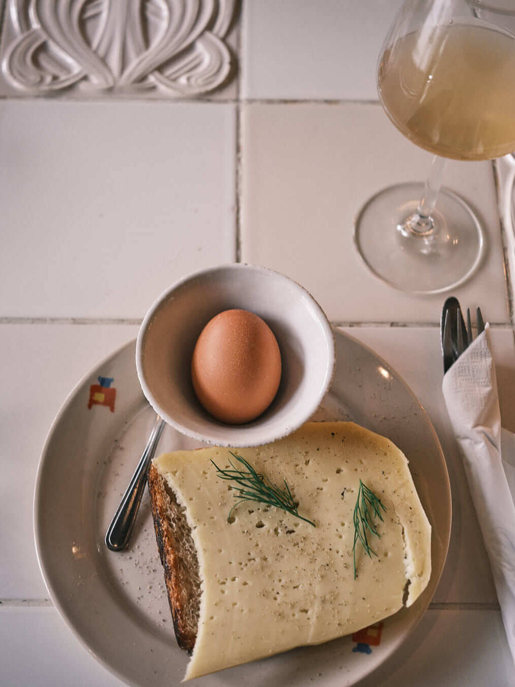 A simple breakfast spread at Material natural wine bar in Berlin, featuring sourdough bread topped with cheese, fresh herbs, and black pepper, served alongside a soft-boiled egg in a ceramic bowl. A comforting and refined start to the day.