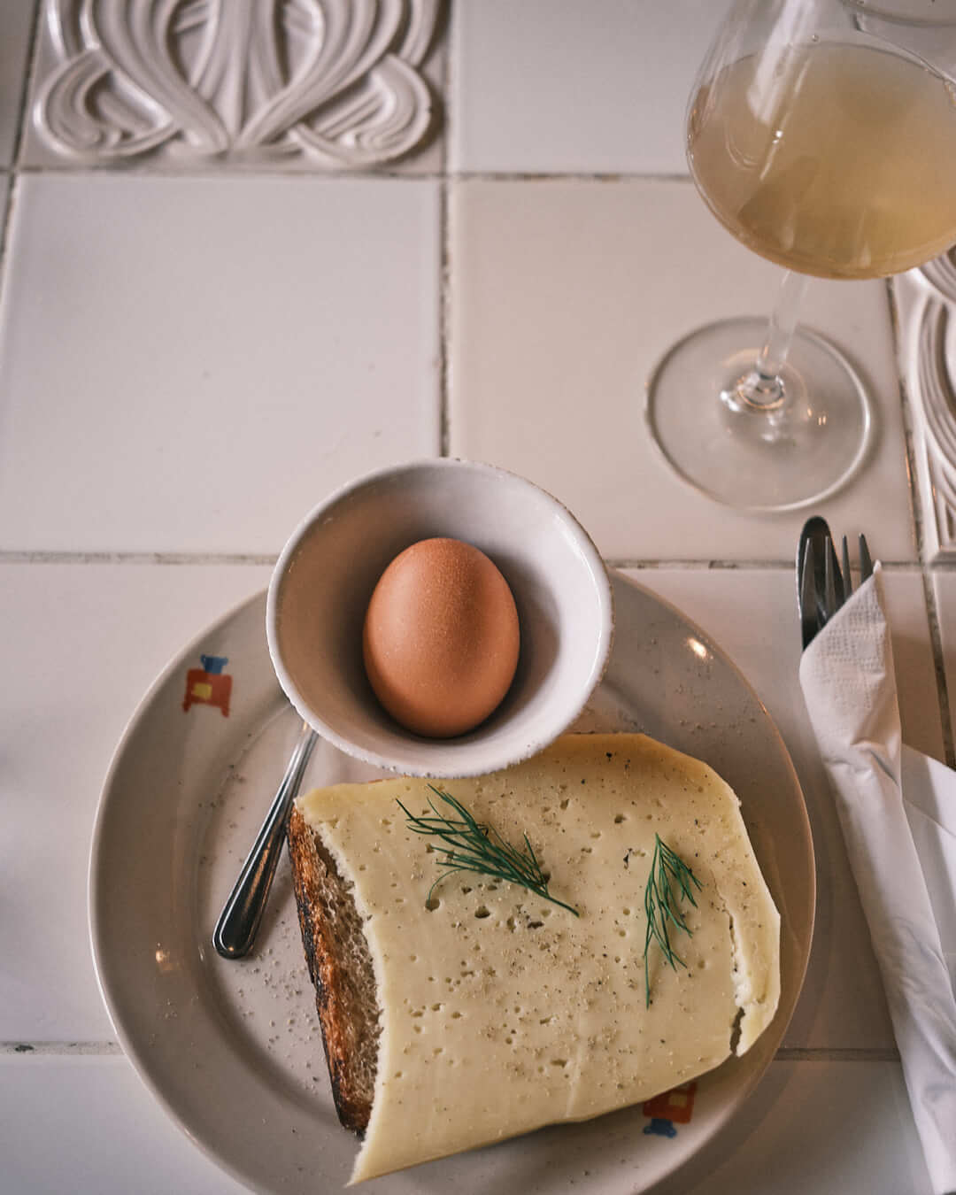 A simple breakfast spread at Material natural wine bar in Berlin, featuring sourdough bread topped with cheese, fresh herbs, and black pepper, served alongside a soft-boiled egg in a ceramic bowl. A comforting and refined start to the day.