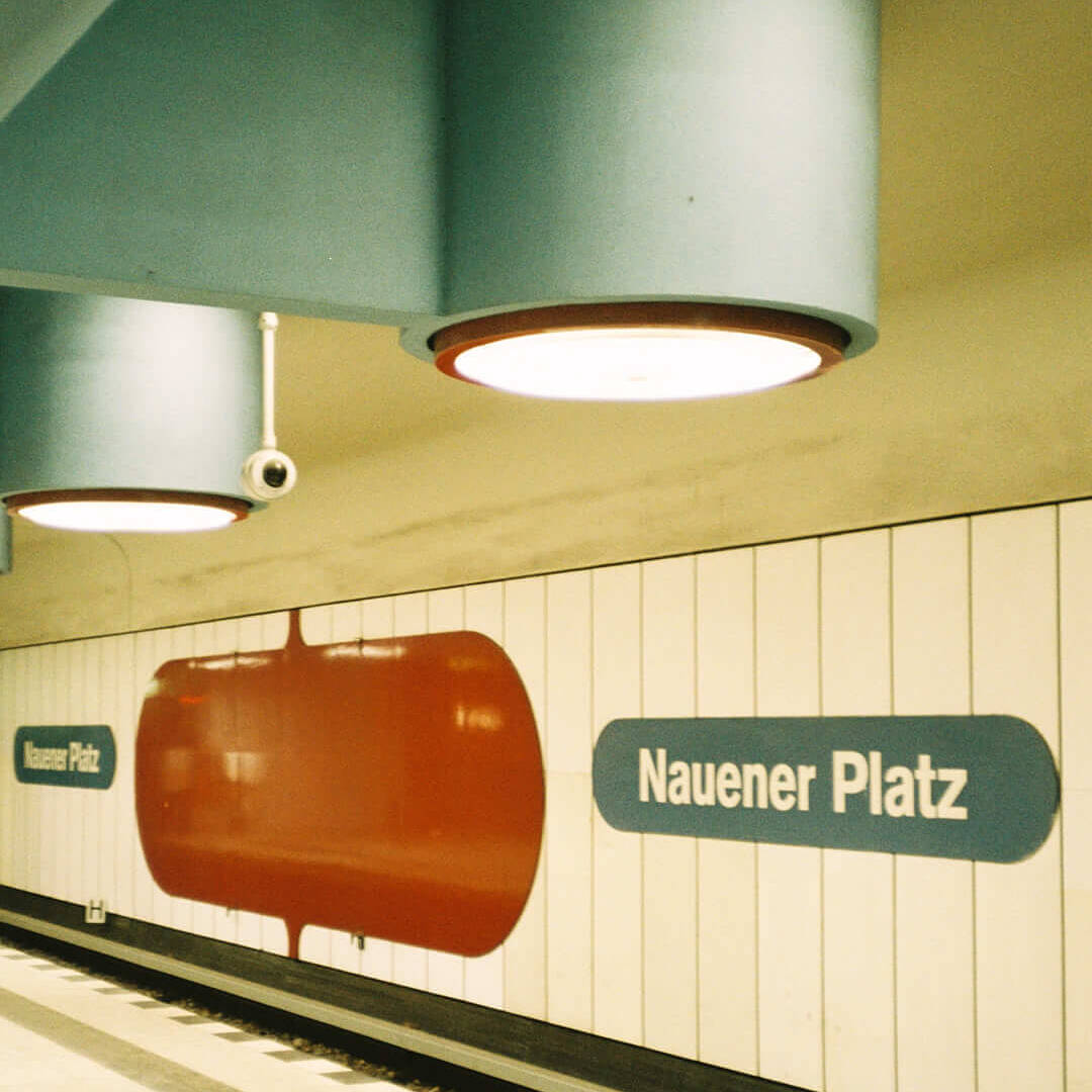 Graphic station wall of Naueners Platz with bold red and blue signage, under round retro ceiling lights.