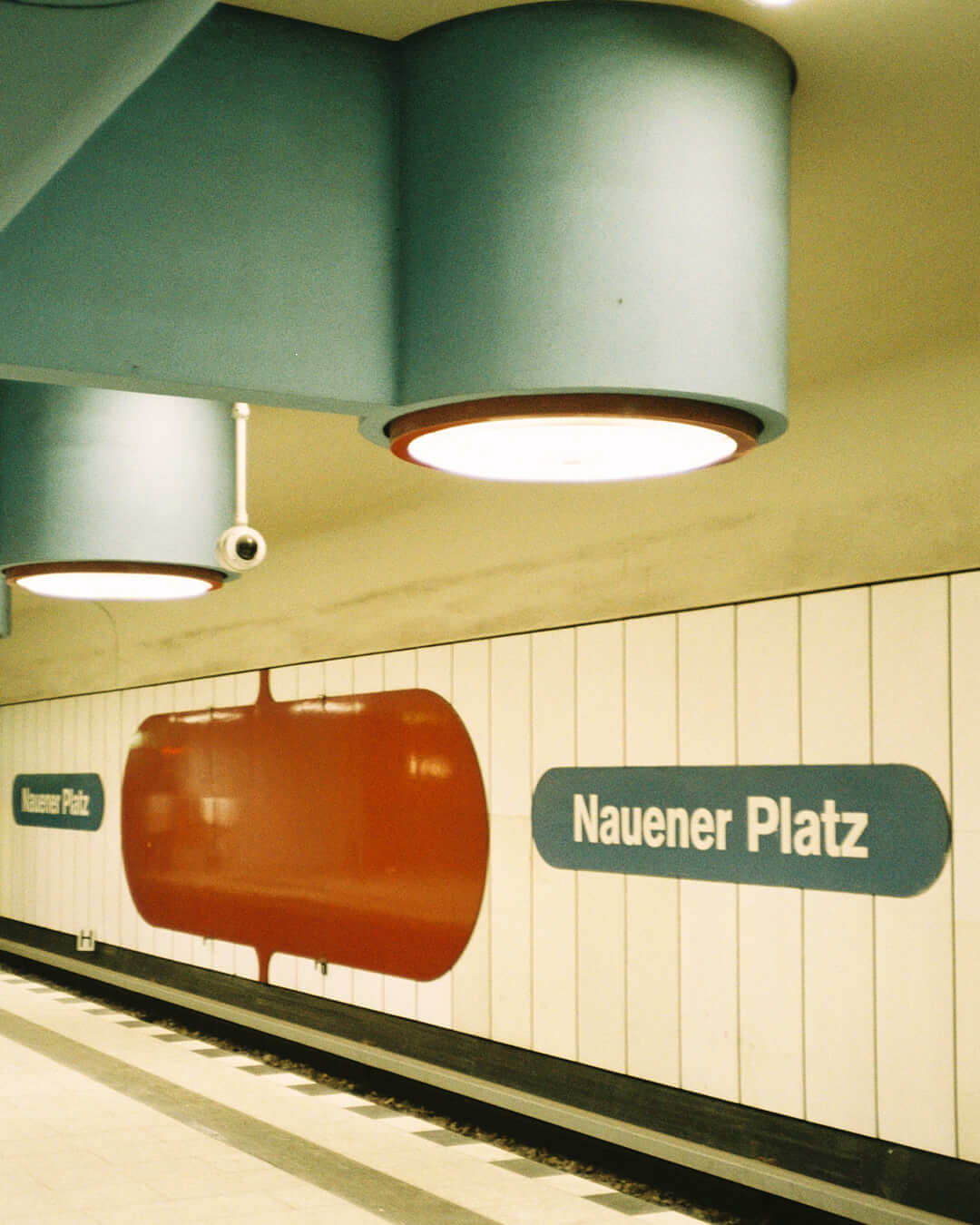 Graphic station wall of Naueners Platz with bold red and blue signage, under round retro ceiling lights.