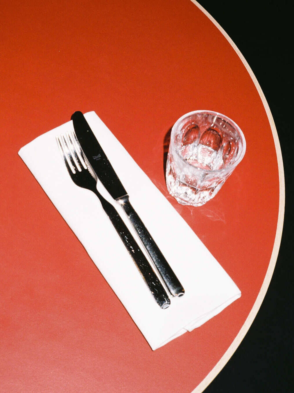 Close-up of a neatly arranged place setting at Trio Berlin, featuring a fork and knife on a white napkin beside a small drinking glass, on a red tabletop.