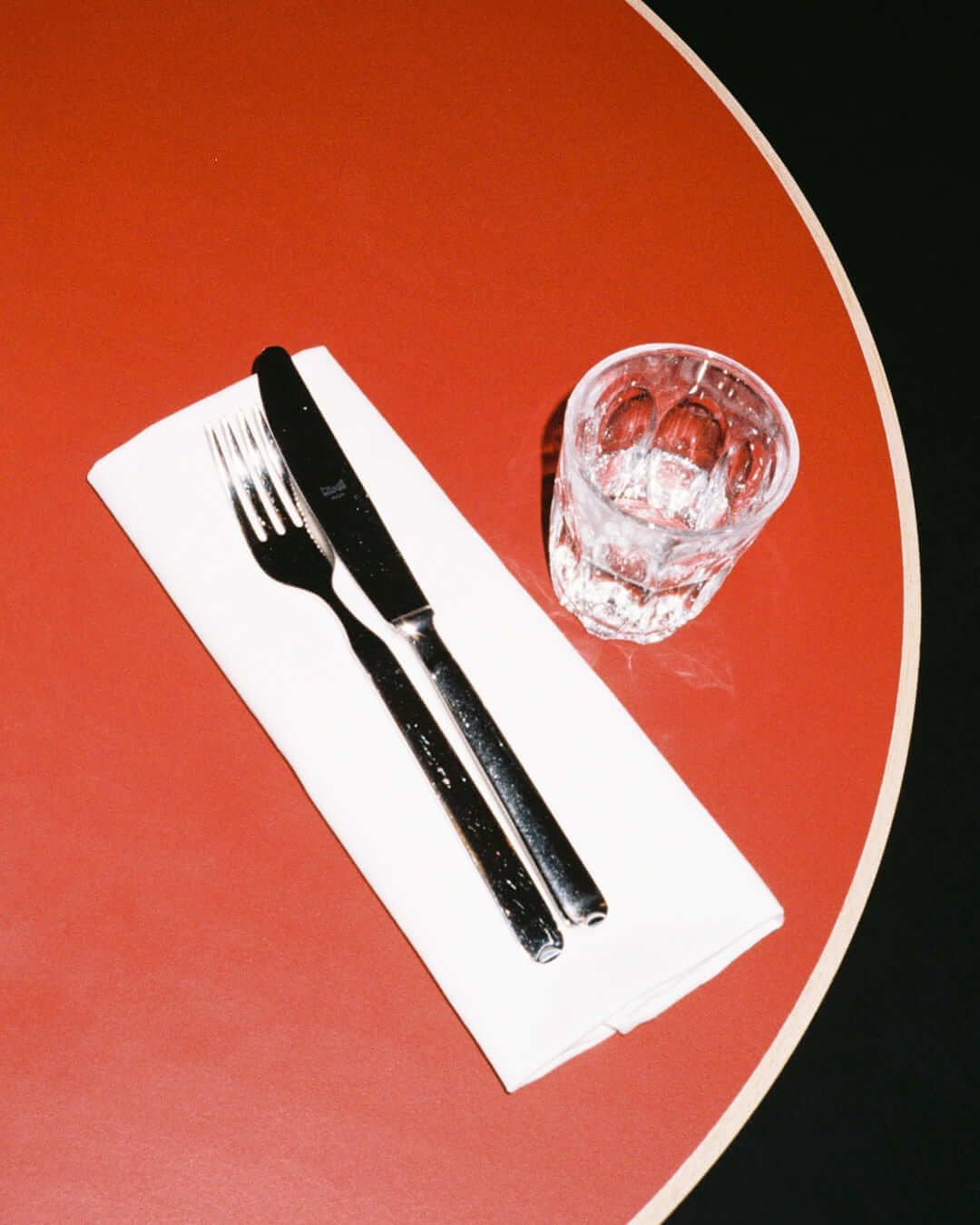 Close-up of a neatly arranged place setting at Trio Berlin, featuring a fork and knife on a white napkin beside a small drinking glass, on a red tabletop.