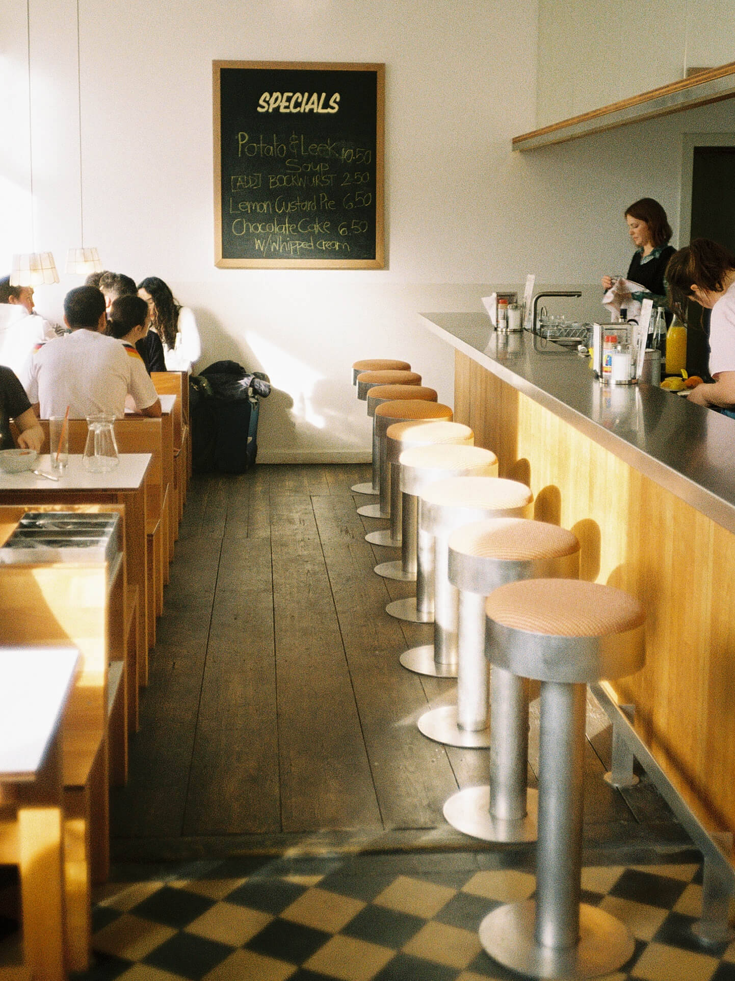 A wide interior view of Onette Berlin featuring a row of sleek retro bar stools along a wooden counter and guests sitting in dining booths. A large chalkboard lists daily specials like Potato & Leek Soup and Lemon Custard Pie. Graphic checkered tile flooring in the foreground.