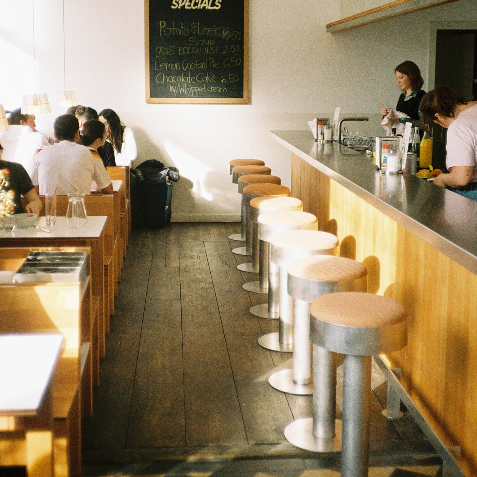 A wide interior view of Onette Berlin featuring a row of sleek retro bar stools along a wooden counter and guests sitting in dining booths. A large chalkboard lists daily specials like Potato & Leek Soup and Lemon Custard Pie. Graphic checkered tile flooring in the foreground.