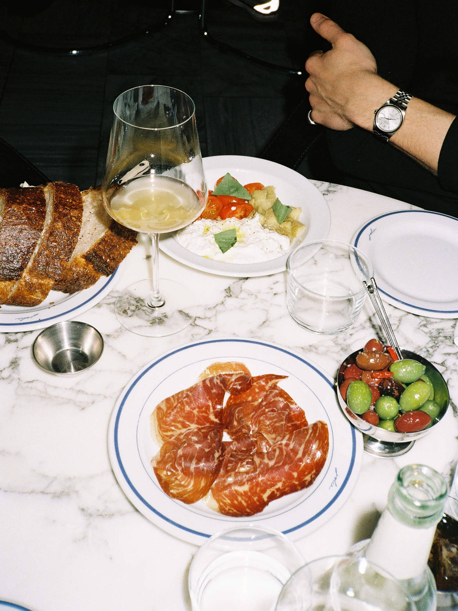 A close-up food photograph of a French charcuterie plate served on a red marble tabletop at Joli Bistrot. The white plate is filled with assorted cured meats, parma ham, and rillettes, garnished with small pickles (cornichons). Shot on 35mm film with direct flash emphasizing the rich food textures.