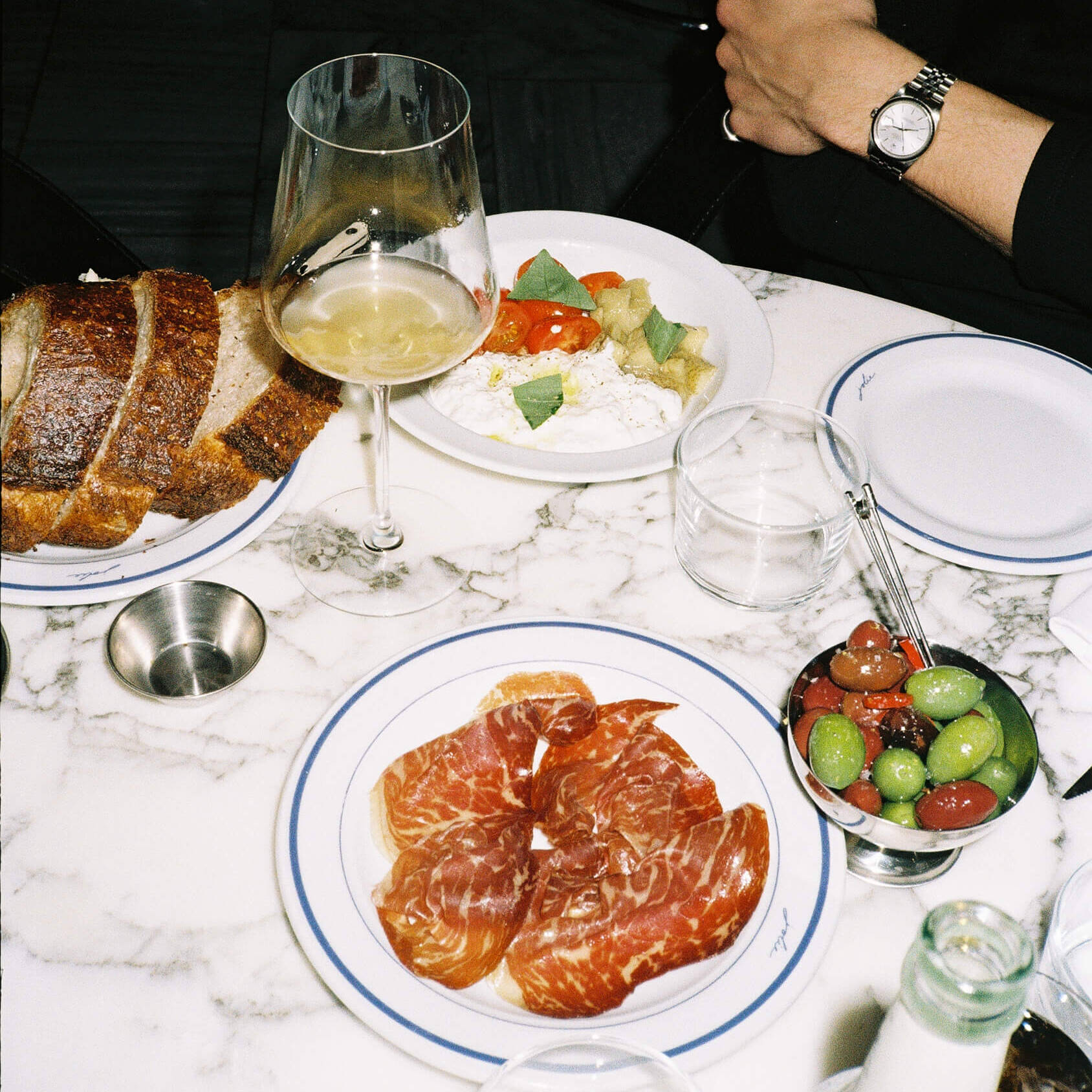 A close-up food photograph of a French charcuterie plate served on a red marble tabletop at Joli Bistrot. The white plate is filled with assorted cured meats, parma ham, and rillettes, garnished with small pickles (cornichons). Shot on 35mm film with direct flash emphasizing the rich food textures.