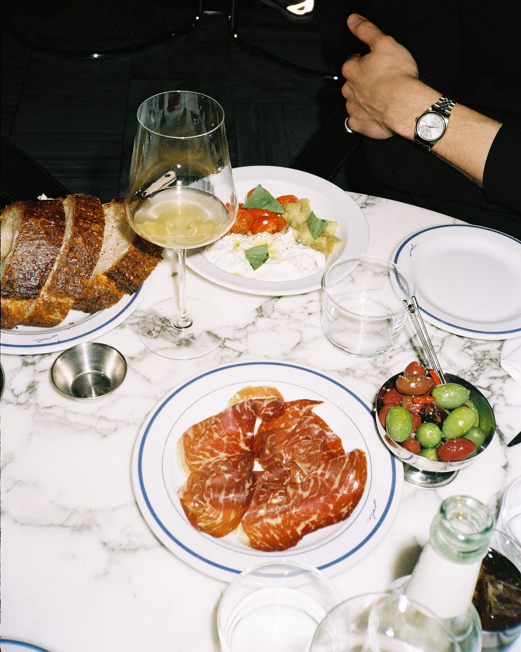 A close-up food photograph of a French charcuterie plate served on a red marble tabletop at Joli Bistrot. The white plate is filled with assorted cured meats, parma ham, and rillettes, garnished with small pickles (cornichons). Shot on 35mm film with direct flash emphasizing the rich food textures.