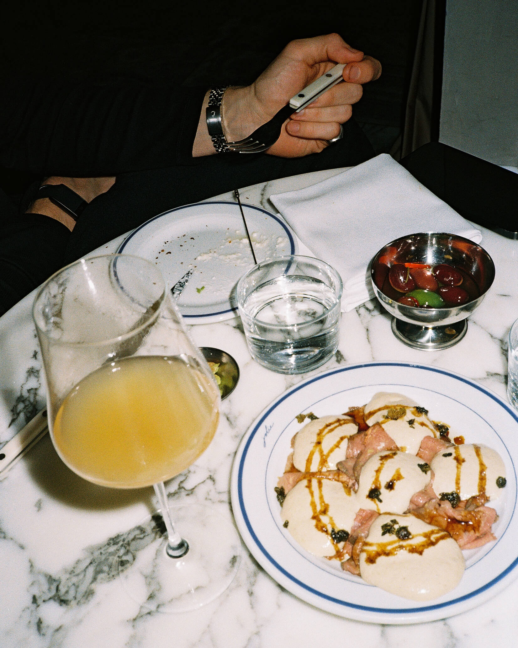 A close-up food photograph taken at Joli Bistrot, featuring a plate of Vitello Tonnato topped with capers and a side of green olives on a red marble table. A person's hand holding a silver fork is poised above the food, and a glass of orange natural wine sits next to the plate. Captured on 35mm analog film with a vivid direct flash.