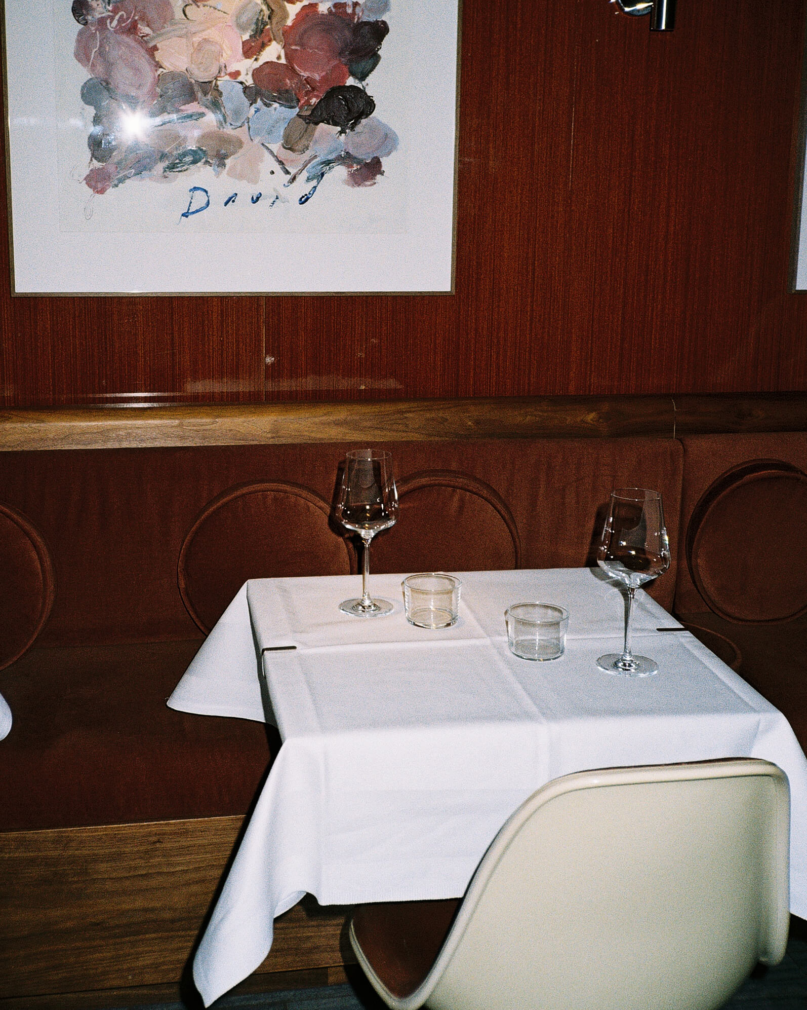An atmospheric interior shot of a dining table beautifully set with a white tablecloth, elegant wine glasses and plates at Joli Bistrot. The entire background features a rich, polished vertical wood paneled wall, reflecting the warm indoor lighting. Shot on 35mm analog film with direct flash.