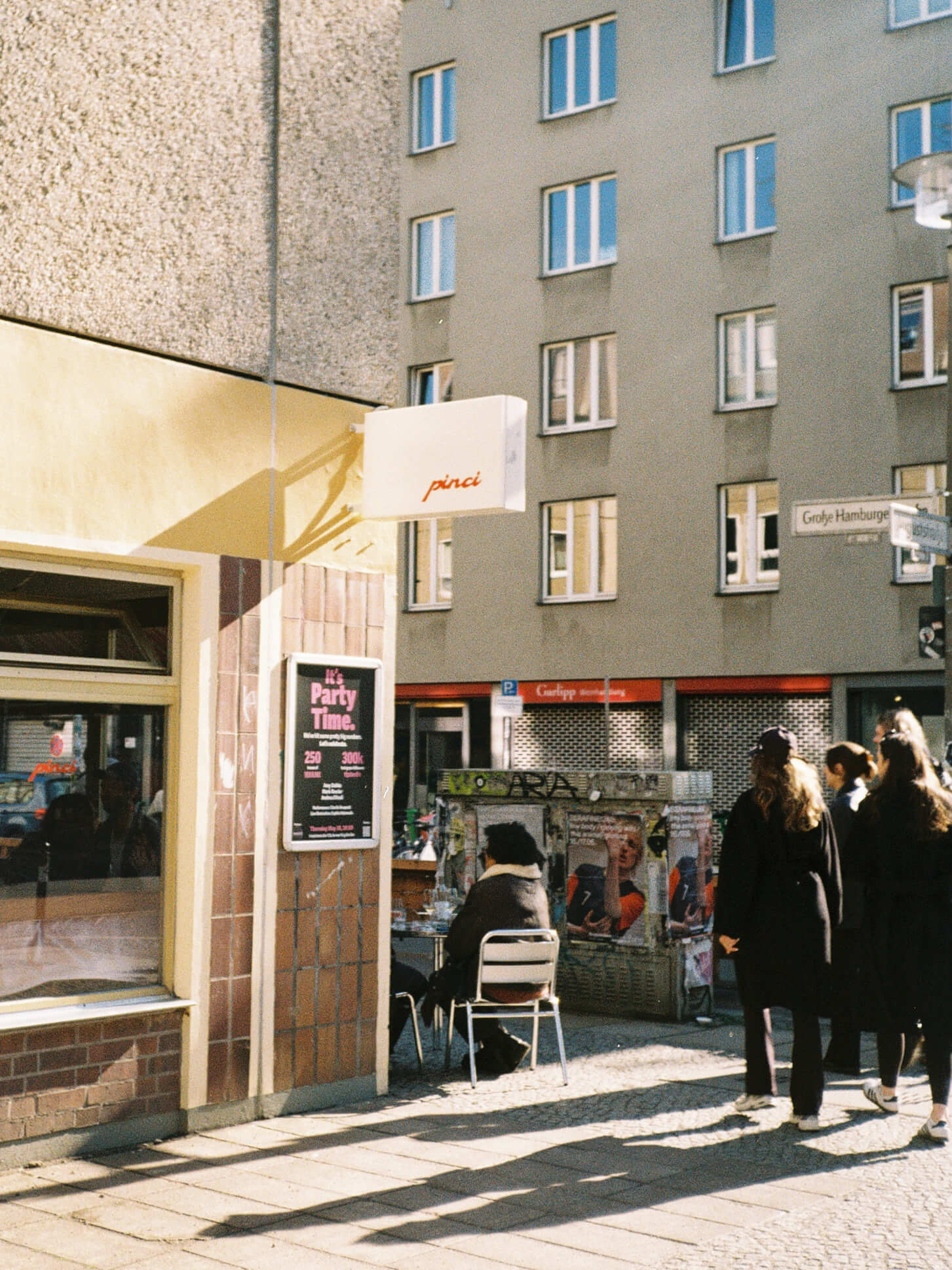 A sunny street corner view of Pinci restaurant in Berlin-Mitte, showing a white minimalist lightbox sign with the red text "pinci" mounted on the wall. A poster on the brown tiled building reads "It's Party Time." Pedestrians are standing on the sidewalk. Analog photography.