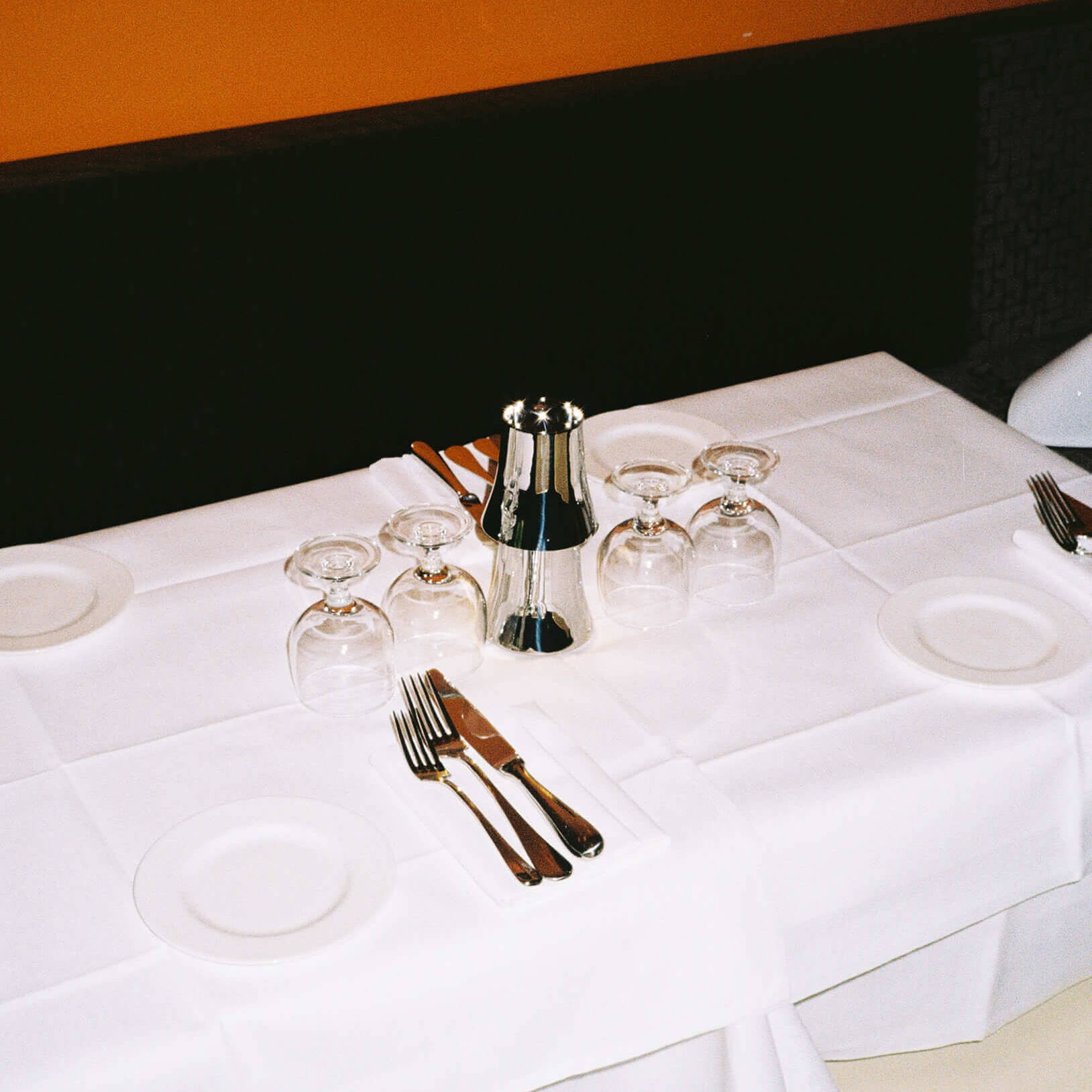 A beautifully set dining table for four at Trattoria Breda under warm lighting, showcasing a crisp white tablecloth, upside down wine glasses, polished silverware, and small white side plates against a dark bench and a solid orange wall background. Analog photography.