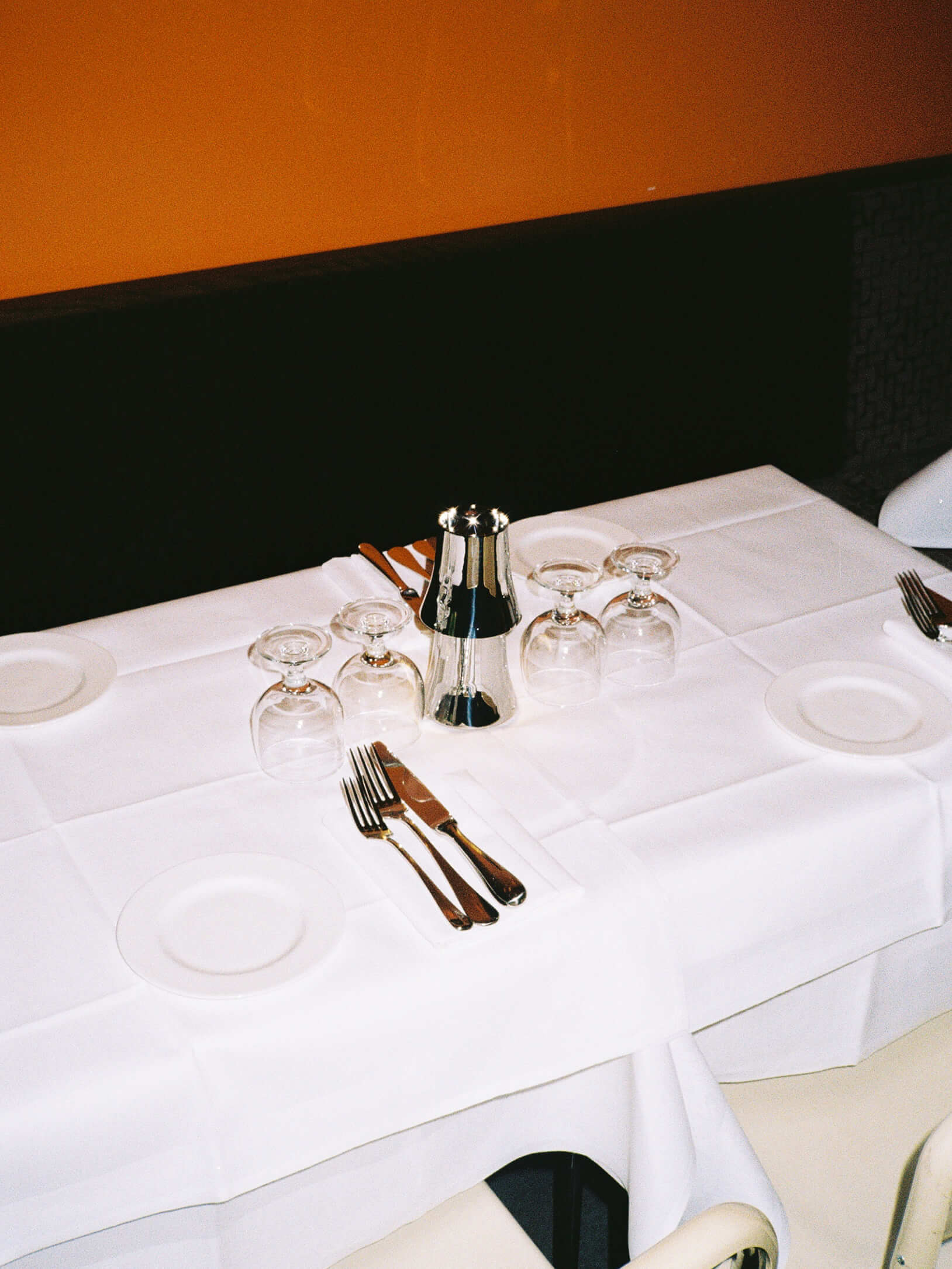 A beautifully set dining table for four at Trattoria Breda under warm lighting, showcasing a crisp white tablecloth, upside down wine glasses, polished silverware, and small white side plates against a dark bench and a solid orange wall background. Analog photography.