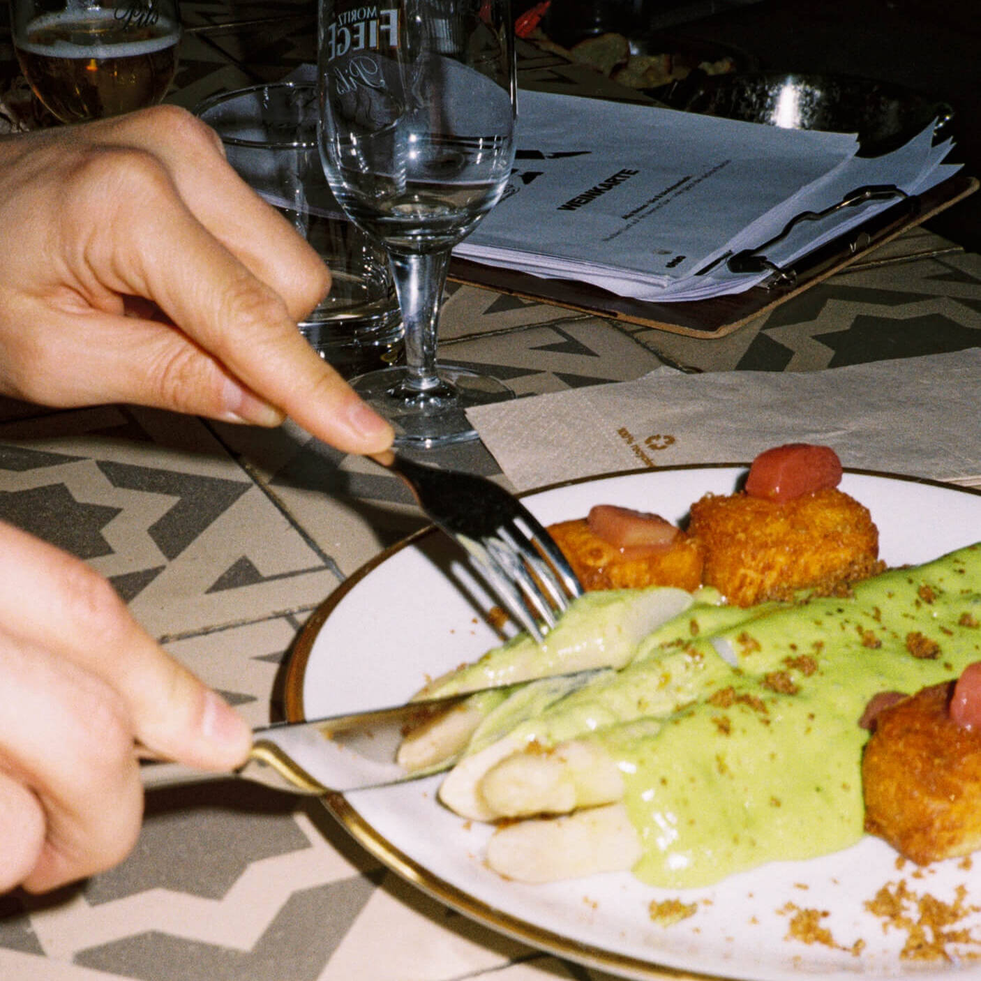 A close-up, action shot of a person’s hands using a knife and fork to cut into white asparagus with green sauce on a plate at Chic Düsseldorf. The background shows water glasses and a clipboard menu. Captured on 35mm film with a direct flash.