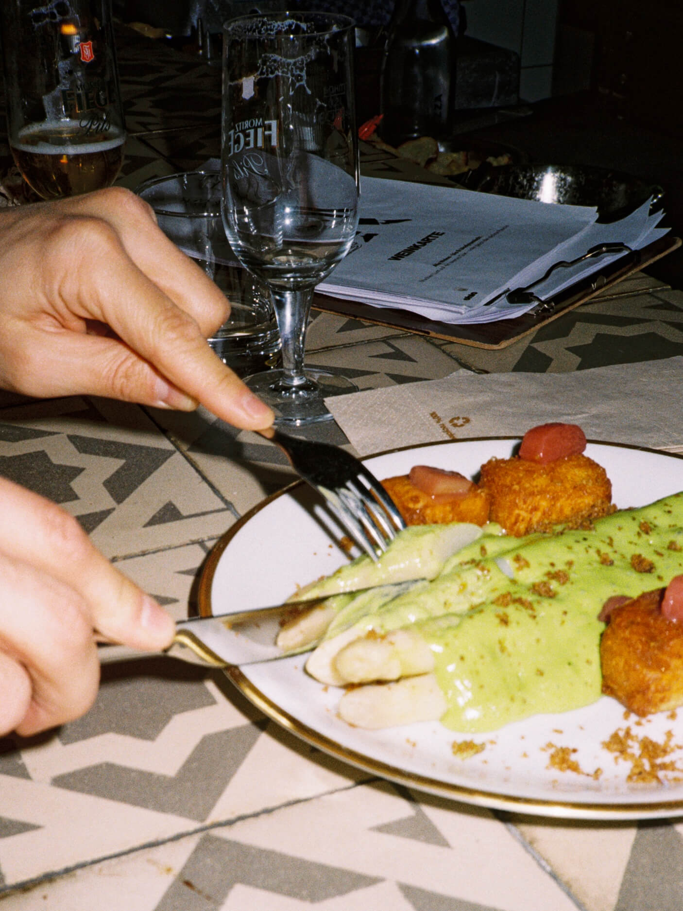 A close-up, action shot of a person’s hands using a knife and fork to cut into white asparagus with green sauce on a plate at Chic Düsseldorf. The background shows water glasses and a clipboard menu. Captured on 35mm film with a direct flash.