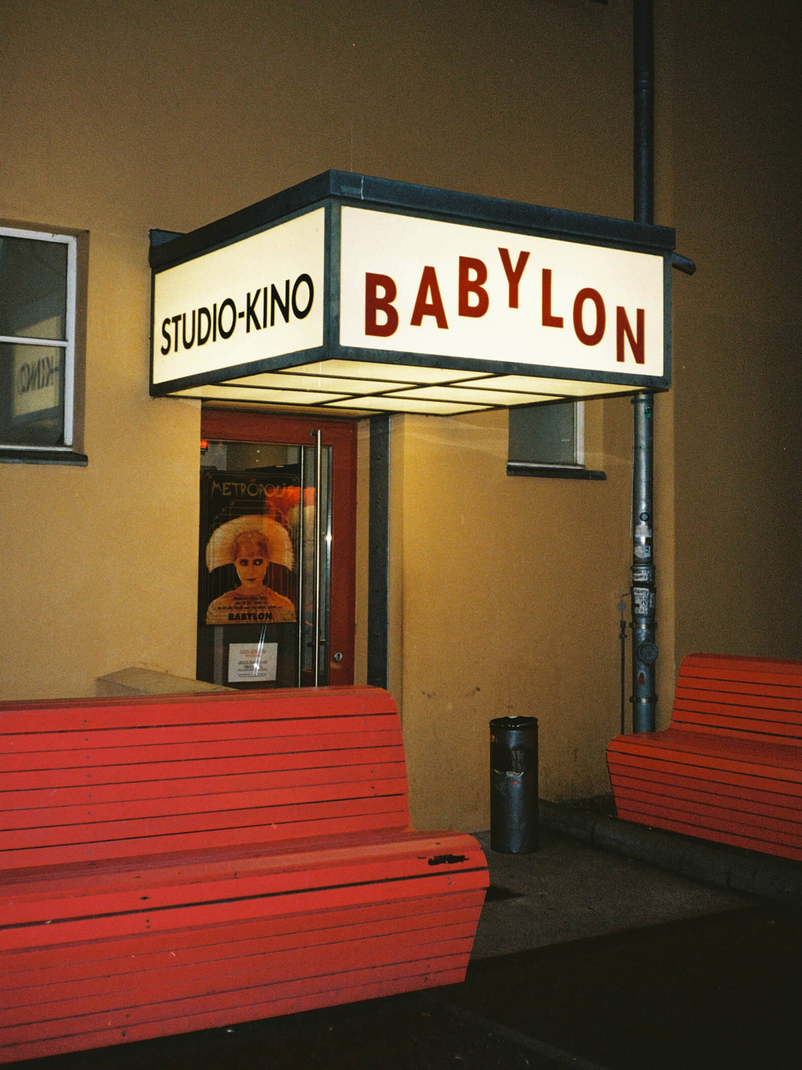 A nighttime photograph showing the side entrance of the Babylon studio cinema under a glowing white lightbox sign reading Studio Kino Babylon, with red wooden benches standing on the pavement. Captured on 35mm film.