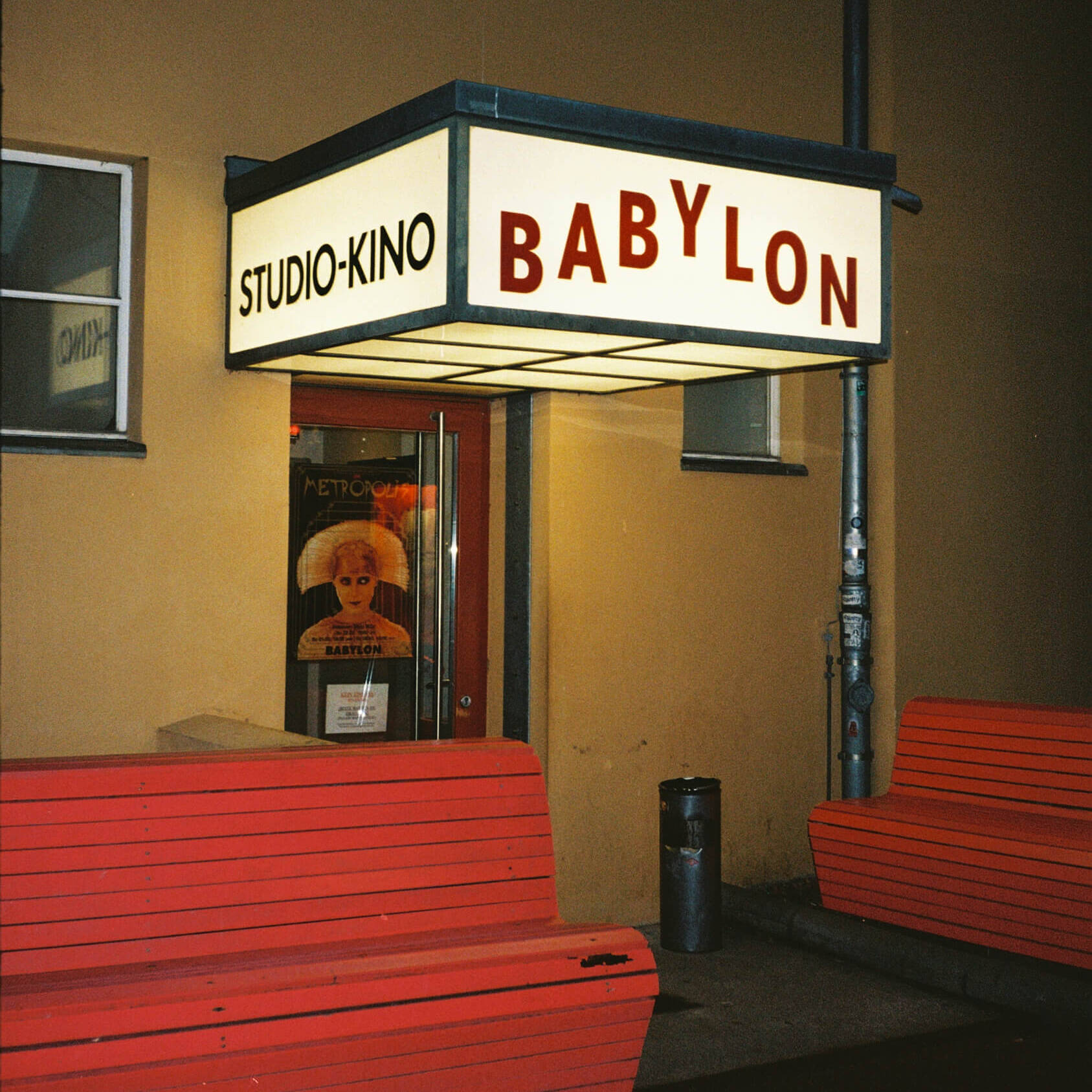 A nighttime photograph showing the side entrance of the Babylon studio cinema under a glowing white lightbox sign reading Studio Kino Babylon, with red wooden benches standing on the pavement. Captured on 35mm film.