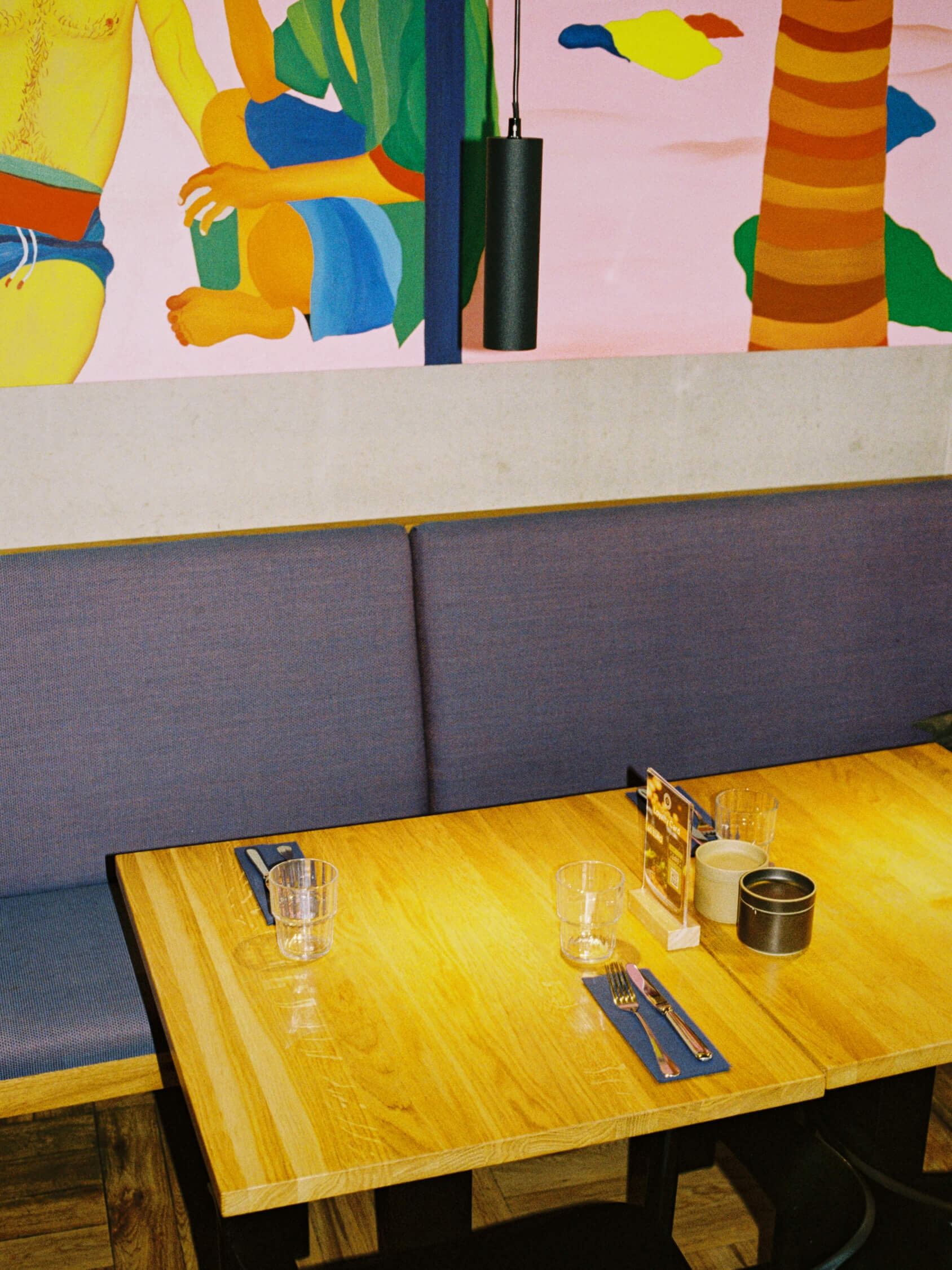 Modern wooden dining table and blue booth seating beneath a colorful beach mural at Frühstück 3000.