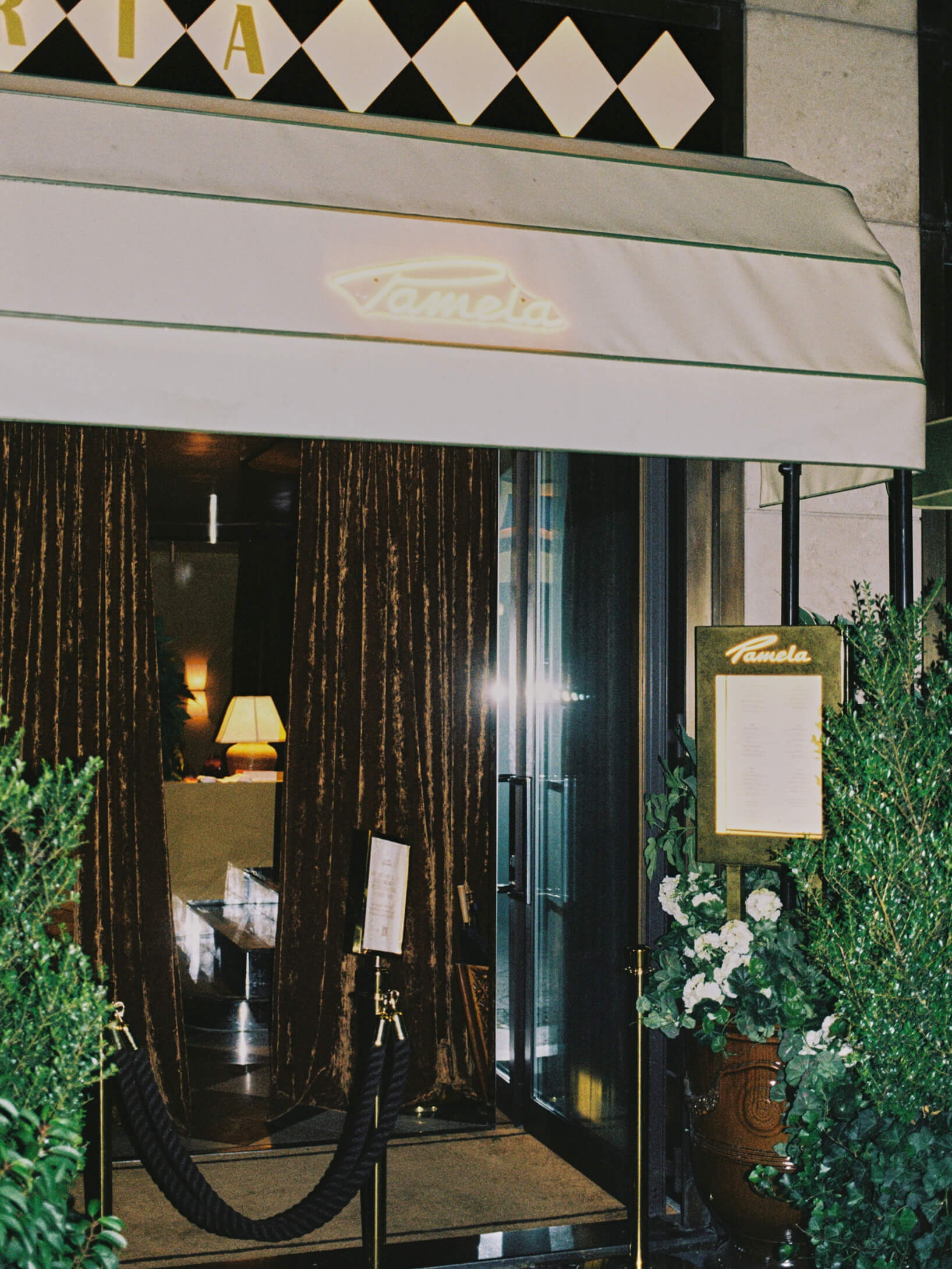 Exterior entrance of Pamela restaurant in Berlin with white awning, neon sign, and velvet curtains at night.