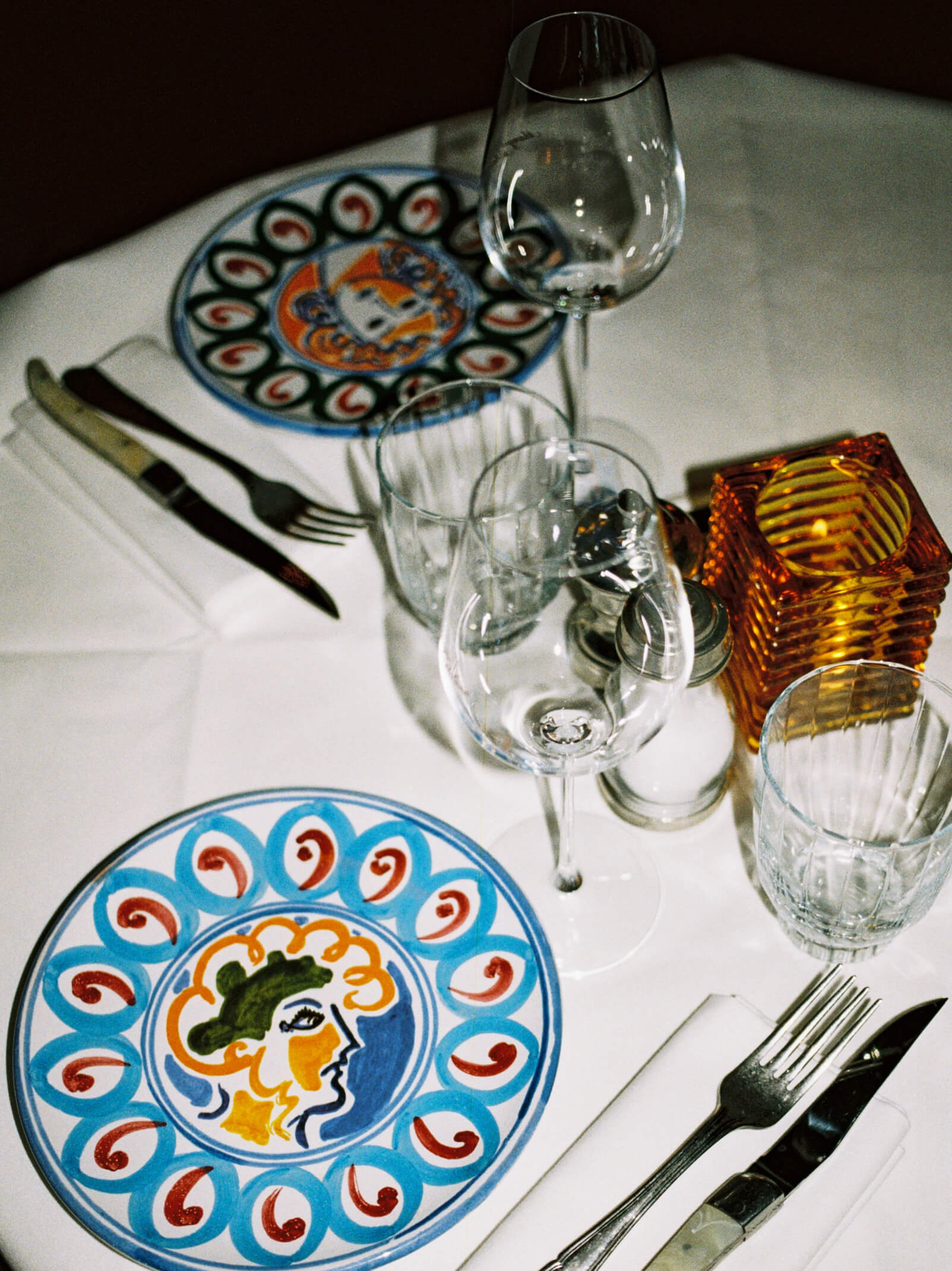 Retro table setting at Pamela Berlin featuring hand-painted Italian ceramic face plates and amber glass candleholder.